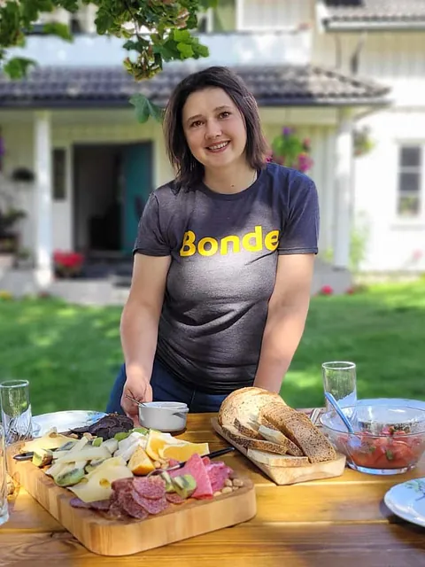 Kvinne i T-skjorte med «Bonde» serverer ost, brød og frukt ute i hage.