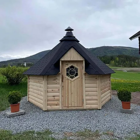 Liten åttekantet grillhytte i tre med svart tak og pyntede blomsterkrukker.