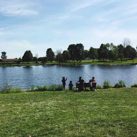 Familie ved innsjøkant fisker og nyter en solrik dag med grønt gress og trær i bakgrunnen.