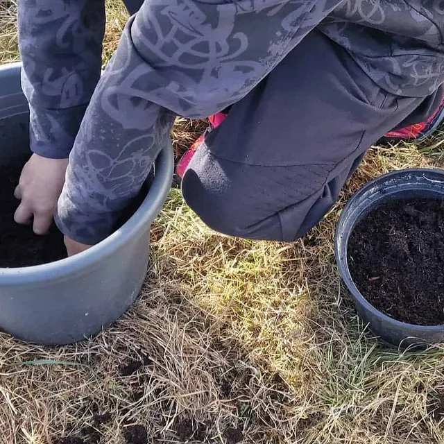 Person planter i stor potte på gress, mindre potte ved siden av.