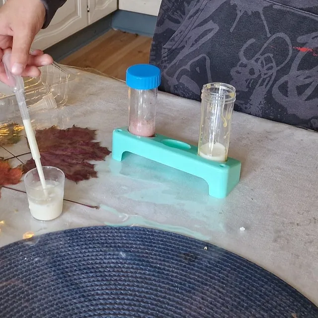 Hånd bruker pipette til å overføre hvit væske til plastbeger ved siden av prøveglass i stativ.