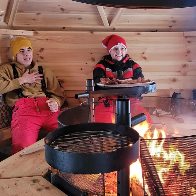 To personer sitter i grillhytte med bål og mat på grillen.